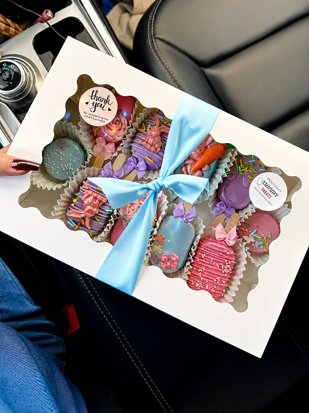 Scalloped window gift box with a blue satin bow holding a spring variety of cakesicles, cookies, and pretzels
